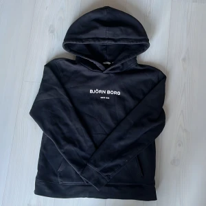 Svart hoodie från Björn Borg - Säljer en stilren svart hoodie från Björn Borg med vit logotyp på bröstet. Tröjan har en klassisk design med huva och en stor ficka framtill. Perfekt för en avslappnad look.
