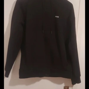 Svart hoodie från Hugo Boss - Säljer en oanvänd svart hoodie från Hugo Boss med diskret logga på bröstet. Perfekt för en avslappnad och trendig look. Hoodien har långa ärmar och dragsko i huvan.