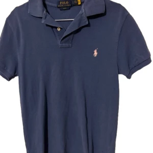 Blå pikétröja från Ralph Lauren - Snygg blå pikétröja från Ralph Lauren med klassisk krage och knappar. Den har en broderad logga på bröstet i rosa. Perfekt att ha i skolan!🔥