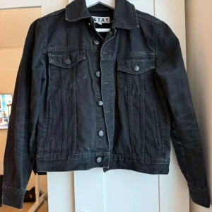 Svart jeansjacka  - Snygg svart jeansjacka från Stay med klassisk design. Jackan har knappar framtill och två bröstfickor med lock. Perfekt för en cool och avslappnad stil. Finns slitage på knapp