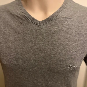 Grå t-shirt från Boss - Snygg grå t-shirt från Boss med v-ringning. Perfekt för en stilren look. Tillverkad i mjukt material för extra komfort. Diskret logga på bröstet.