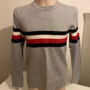 Grå tröja från Tommy Hilfiger - Säljer en stilren grå tröja från Tommy Hilfiger med svarta, vita och röda ränder över bröstet och ärmarna. Tröjan har långa ärmar och en rund halsringning. Perfekt för en avslappnad och snygg look. Liten i storlek. Är mer som stl. M