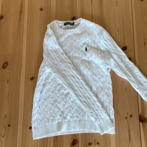 Vit kabelstickad tröja från Ralph Lauren - Säljer en klassisk vit kabelstickad tröja från Ralph Lauren med det ikoniska broderade logotypen på bröstet. Tröjan har långa ärmar och ribbade muddar vid ärmslut och nederkant. Perfekt för en stilren look! Perfekt skick och inga hål eller defekter, om du är intresserad är det bara att höra av sig. Priset är inte hugget ur sten.