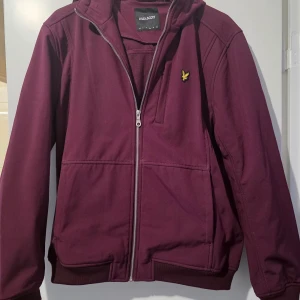 Vinröd jacka från Lyle & Scott oanvänd - Snygg vinröd jacka från Lyle & Scott med dragkedja och huva. Jackan har en broderad logotyp på bröstet och är tillverkad i ett mjukt material. Fodrad insida och helt oanvänd