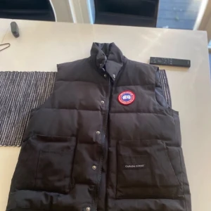 Svart dunväst från Canada Goose - Säljer en svart dunväst från Canada Goose med knappar och dragkedja. Västen har två stora fickor framtill och en broderad logotyp på bröstet. Perfekt för kyliga dagar!