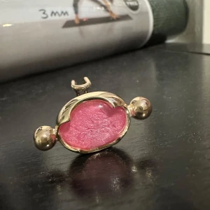 Guldpläterad ring med rosa detalj från Ioaku - Elegant guldpl ring med en iögonfallande rosa detalj i mitten. Ringen har en unik design med två kulor på sidorna och en justerbar bandstruktur. Perfekt för att ge en färgklick till din outfit.
