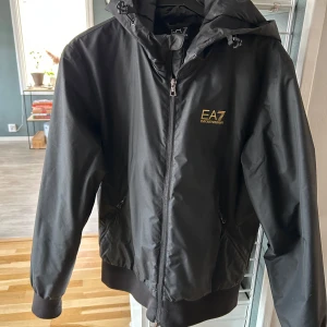 Svart vindjacka från EA7 - Snygg svart vindjacka med lättare vaddering från EA7 med dragkedja och huva. Jackan har ribbade muddar och EA7-logga i guld på bröstet. Passar både vår och sommar