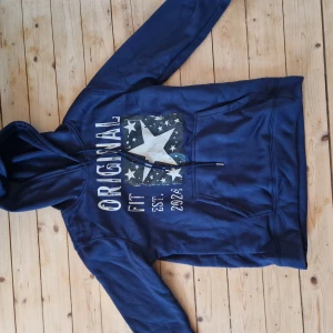 Original hoddie - Säljer en blå hoodie med tryck av en stor stjärna och texten 'Original Fit Est. 2024'. Hoodien har en justerbar huva med snören och en stor ficka framtill. Perfekt för en avslappnad stil.