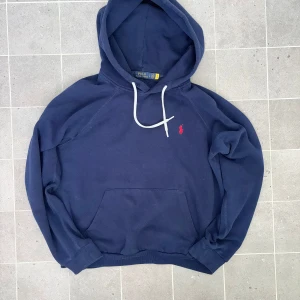 Mörkblå hoodie från Polo Ralph Lauren - Säljer en snygg mörkblå hoodie från Polo Ralph Lauren med den klassiska röda loggan på bröstet. Tröjan har en justerbar huva med vita snören och en praktisk magficka. Perfekt för en avslappnad stil.storlek L barn.ETT LITET HÅLL NEDANFÖR DÄR SNÖRET KOMMER UT SE SISTA BILDEN