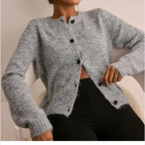 Grå stickad cardigan från Nelly - Mysig grå kofta med knappar framtill. Den har en normal passform och är tillverkad i ett mjukt material som ger en skön känsla. Perfekt för kyligare dagar.