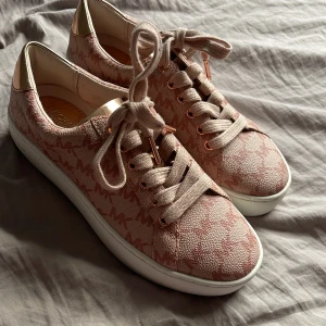 Rosa sneakers från Michael Kors - Snygga rosa sneakers från Michael Kors med ett diskret mönster och snörning. Skorna har en vit sula och detaljer i guld. Perfekta för en stilren look. Aldrig använda