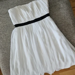 Vit axelbandslös klänning med svart band - Säljer en elegant vit axelbandslös klänning i korsett modell med ett svart band i midjan. Klänningen har en luftig och flödande design, perfekt för sommarens festligheter. 