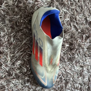 Fotbollsskor från Adidas - Snygga fotbollsskor från Adidas i vitt med röda och blå detaljer. Skorna har en modern design utan snörning och är perfekta för snabba rörelser på planen.