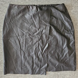 Svart kjol i syntetmaterial - Elegant svart kjol i syntetmaterial med en stilren design. Kjolen har en rak passform och en diskret slits framtill som ger en modern touch. Perfekt för en chic och minimalistisk look.