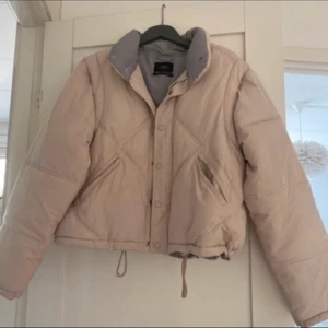 Beige pufferjacka med avtagbara ärmar - Snygg beige pufferjacka med avtagbara ärmar som gör den till en väst. Jackan har knappar och dragsko i nederkant för justerbar passform. Perfekt för kyliga dagar när du vill ha lite extra värme.