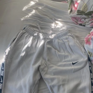 Vita mjukisbyxor från Nike - Säljer ett par vita mjukisbyxor från Nike med elastisk midja och snörning. Byxorna har sidoremsor med nike loggan och säljs för att dom är för små