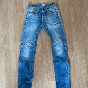 Blå jeans från Replay - Säljer ett par snygga blå jeans från Replay med en klassisk femficksdesign och slitna detaljer. Jeansen har en knappgylf och är perfekta för en avslappnad stil. Märkesdetaljer syns på baksidan och insidan av midjan.