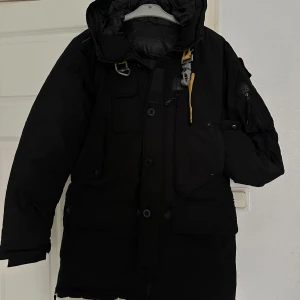 Svart parkajacka med huva - Snygg svart parkajacka med huva och flera praktiska fickor. Jackan har både dragkedja och knappar framtill samt en cool detalj med gul snörning vid kragen. Perfekt för kyliga dagar.