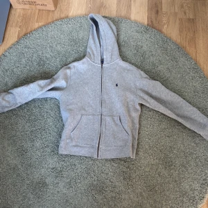 Grå hoodie från Polo Ralph Lauren - Säljer en stilren grå hoodie från Polo Ralph Lauren med dragkedja och kängurufickor. Den har en broderad logotyp på bröstet och är perfekt för en avslappnad look. Huvan ger extra komfort och stil.skicket är 9/10 inga skador men skulle inte säga 10/10 fel att använd några få tal gånger
