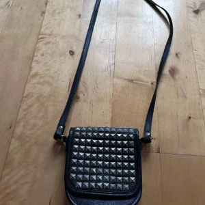Svart axelväska med nitar - Säljer en supercool svart axelväska med silverfärgade nitar på framsidan. Väskan är perfekt för att ge din outfit en edgy touch. Den har en lång rem så du kan bära den bekvämt över axeln. Passar perfekt till både vardag och fest!