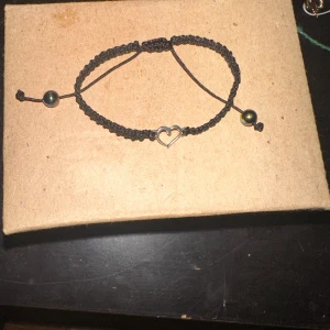Svart armband med hjärtdetalj - Säljer ett snyggt svart armband med en söt hjärtdetalj i mitten. Armbandet är justerbart och har små pärlor vid ändarna. Perfekt för att ge en enkel men stilren look till din outfit!
