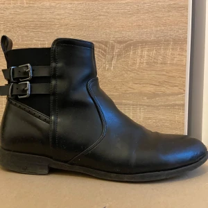 Svarta skinnstövletter med spännen - Säljer ett par snygga svarta stövletter i skinn. De har två dekorativa spännen på sidan och en praktisk dragkedja för enkel på- och avtagning. Perfekta för både vardag och fest!