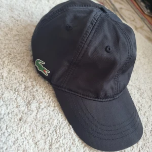 Svart keps från Lacoste - Säljer en snygg svart keps från Lacoste.. Kepsen är i polyester och har en sportig stil. I bra skick