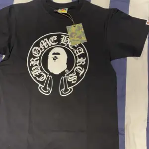 Säljer en svart t-shirt från det coola samarbetet mellan Chrome Hearts och A Bathing Ape. T-shirten har ett stort tryck med 'Chrome Hearts' och 'Baby Milo' i vitt på både fram- och baksidan. Den är kortärmad och perfekt för en avslappnad stil. Taggen är i camo-mönster vilket ger en extra touch.