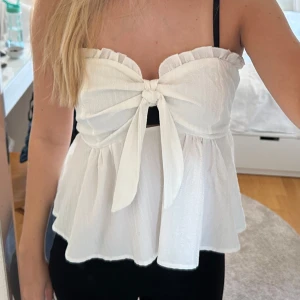 Vit axelbandslös topp med knyt - Säljer en superfin vit axelbandslös topp med knyt framtill. Den har en söt volangdetalj upptill och är lite croppad, vilket gör den perfekt för sommaren. Toppen är i ett lätt och luftigt material, perfekt för varma dagar!