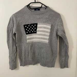 Grå tröja med flaggmotiv från GANT - Säljer en stilren grå tröja från GANT med ett stort flaggmotiv på framsidan. Tröjan har långa ärmar och ribbade kanter vid ärmslut och nederkant. Perfekt för en avslappnad stil.