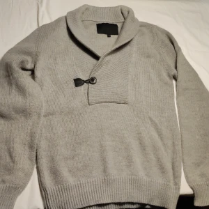 Grå stickad tröja med knappdetalj - Säljer en stilren grå stickad tröja med en unik knappdetalj vid halsen. Tröjan har långa ärmar och ribbade muddar vid ärmslut och nederkant. Perfekt för kyliga dagar!