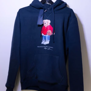 Mörkblå hoodie från Polo Ralph Lauren - Helt ny mörkblå hoodie från Polo Ralph Lauren med en ikonisk nallebrodyr på framsidan. Tröjan har en klassisk känguruficka och justerbar huva med dragsko. Perfekt för en avslappnad stil.