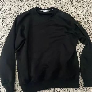 Svart sweatshirt från Stone Island - Säljer en stilren svart sweatshirt från Stone Island. Tröjan har långa ärmar och en rund halsringning. Den ikoniska Stone Island-loggan sitter på ärmen. Perfekt för en casual look.