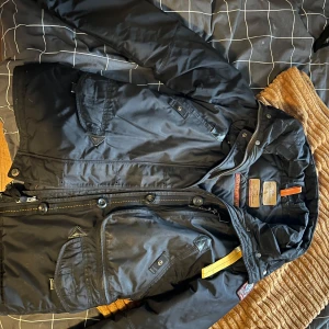 Svart jacka från Parajumpers - Säljer en svart jacka från Parajumpers Masterpiece Series. Jackan har en huva med pälsdetaljer och flera praktiska fickor. Den har både dragkedja och knappar framtill samt en gul remdetalj vid kragen. Perfekt för kyligare väder! 