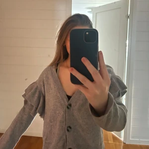 Grå kofta med volangdetaljer - Säljer en mysig grå kofta med volangdetaljer på ärmarna. Koftan har knappar framtill och är perfekt för kyligare dagar. 