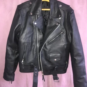 Svart skinnjacka från Cudeta - Snygg svart skinnjacka från Cudeta med klassisk bikerstil. Jackan har dragkedja, knappar och ett bälte i midjan. Den är quiltad på insidan för extra komfort. Perfekt för en cool look! Oanvänd!