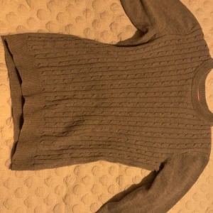 Grå stickad tröja från Mywear Woman - Säljer en stilren grå stickad tröja från Mywear Woman. Tröjan har ett klassiskt flätmönster och långa ärmar, perfekt för kyligare dagar. Den är mjuk och bekväm, idealisk för både vardag och avslappnade tillfällen.