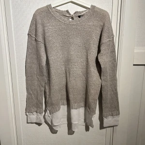 Beige tröja med vit detalj från must have knit collection  - Säljer en beige tröja med en vit underdel som ger en lager-på-lager-look. Tröjan har långa ärmar och en rund halsringning. Perfekt för en avslappnad stil.