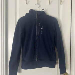 Mörkblå jacka från Sail Racing - Snygg mörkblå jacka från Sail Racing med dragkedja och huva. Perfekt för kyliga dagar. Jackan har en praktisk bröstficka med dragkedja och är tillverkad i ett mjukt material. OBS fickan på vid bröstkorgen sitter löst.
