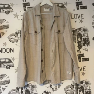 Beige overshirt  - Snygg beige overshirt  med långa ärmar och knappar framtill. Perfekt för lager-på-lager-stil. Den har en klassisk krage och två bröstfickor med lock. En stilren och mångsidig skjorta för alla tillfällen. Aldrig använd