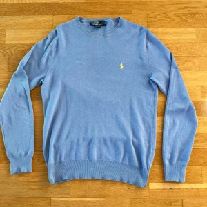 Ljusblå tröja från Ralph Lauren - Säljer en stilren ljusblå tröja från Ralph Lauren med det klassiska gula logotypbroderiet på bröstet. Tröjan har långa ärmar och ribbade muddar vid ärmslut och nederkant. Perfekt för en avslappnad men snygg look. Nypris: 1499