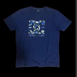 Blå t-shirt från DC  - Fet blå DC T-shirt med DC loggan i  kamouflagemönster på bröstet. Sällan använd, pris kan diskuteras, skriv vid funderingar.