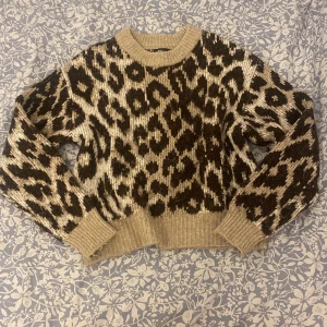 Leopardmönstrad stickad tröja från Gina Tricot - Säljer en snygg leopardmönstrad stickad tröja från Gina Tricot. Tröjan har en beige bas med mörkbruna fläckar och långa ärmar. Perfekt för att ge din outfit en trendig touch. 🐆