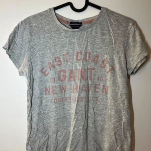 Grå t-shirt från Gant - Säljer en grå t-shirt från Gant med tryck i rosa som säger 'East Coast New Haven'. T-shirten har en rund hals och korta ärmar. 