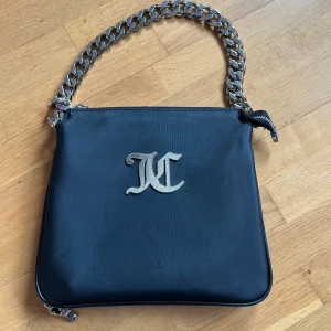 Svart axelväska med kedja från Juicy couture  - Snygg svart axelväska med en kraftig silverkedja som axelrem. Väskan har en stilren design med ett metalliskt emblem på framsidan och dragkedja runtom. Perfekt för att ge en edgy touch till din outfit. Pågrund av dragkedjan ner till på väskan kan man göra den större och mindre. 