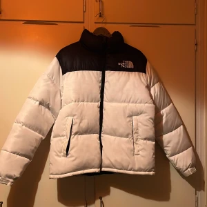 Vit och svart dunjacka från The North Face - Säljer en snygg vit och svart dunjacka från The North Face. Jackan har en dragkedja framtill och två praktiska fickor. Perfekt för kalla dagar med sin pufferdesign och långa ärmar. Logan är synlig på bröstet och baksidan.