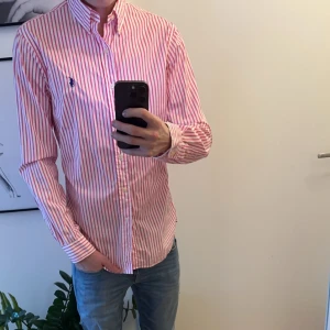 Rosa - och vitrandig skjorta från Ralph Lauren - Snygg röd- och vitrandig skjorta från Ralph Lauren med klassisk krage och knappar framtill. Aldrig använd. Säljs för den bara hänger i min garderob. Storlek M 