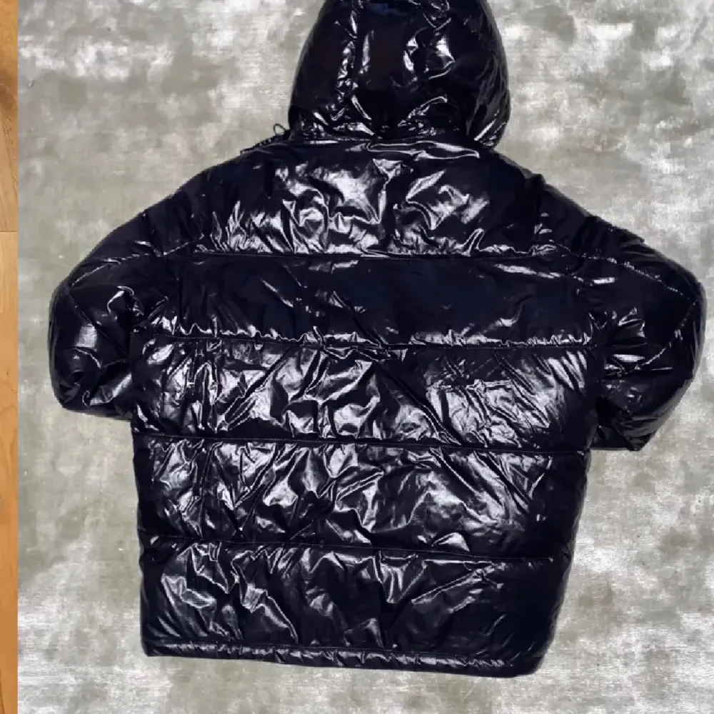 Snygg svart pufferjacka från Michael Kors med glansig yta och huva. Jackan har en dragkedja framtill och är quiltad för extra värme. Perfekt för kyliga dagar.. Takit.