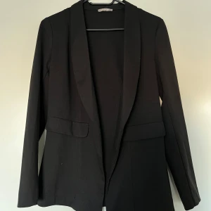 Kavaj, Orsay, 36, svart - Svart kavaj från Orsay i storlek 36. Kavajen är i nyskick.  Normal passform. Elastisk. Utan innertyg. 2 låtsasframfickor fram. 73% polyester, 22% viskos, 5% elastan.   Mått:  Längd ca 72 cm Byst ca 44 cm Midja ca 43 cm Ärmlängd mätt från axel ca 60 cm