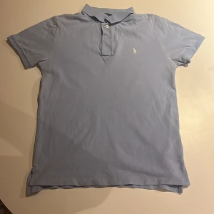 Ljusblå pikétröja från Ralph Lauren - Säljer en stilren ljusblå pikétröja från Ralph Lauren med klassisk krage och knappslå. Tröjan har en broderad logotyp på bröstet och är perfekt för en avslappnad men snygg look. Passar bra till både jeans och chinos. Storlek L i barn